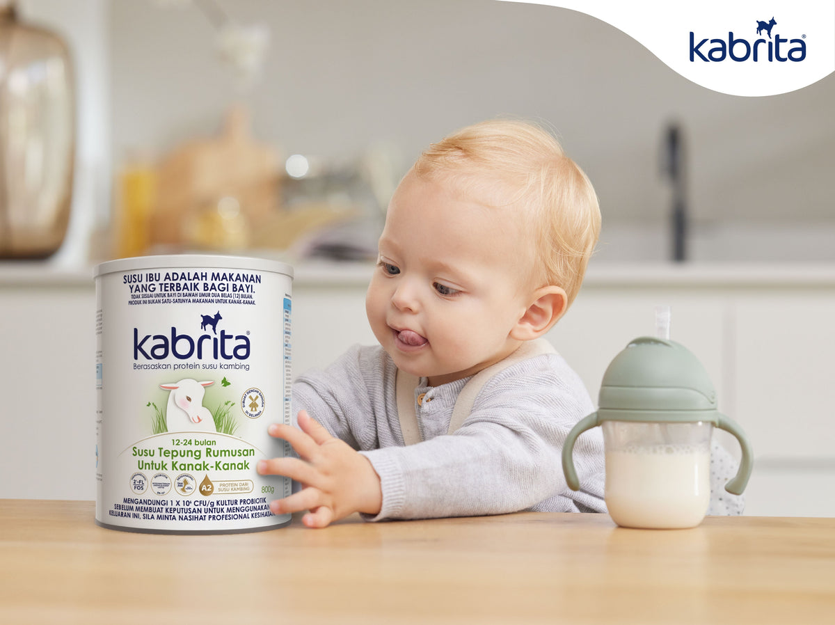 Child beside a Kabrita goat milk formula tin and a handled cup on a table.Kanak-kanak di sebelah tin rumusan susu kambing Kabrita dan cawan berpemegang di atas meja.
