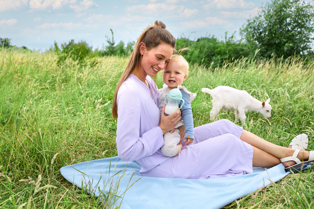 Mother sitting with her child and a cup in a meadow with white goats nearby.
Ibu duduk bersama anak dan cawan di padang rumput dengan kambing putih berdekatan.